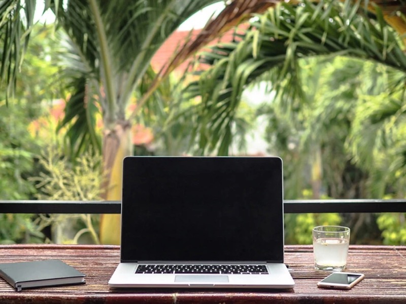 Palmiyelerin önündeki balkon masasında laptop, defter ve bir bardak su